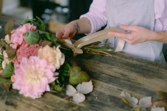 Hände arrangieren zarte rosa und weiße Blüten in rustikalem Holzcontainer - Blumenbinde-Workshop Kreativ & Blüte